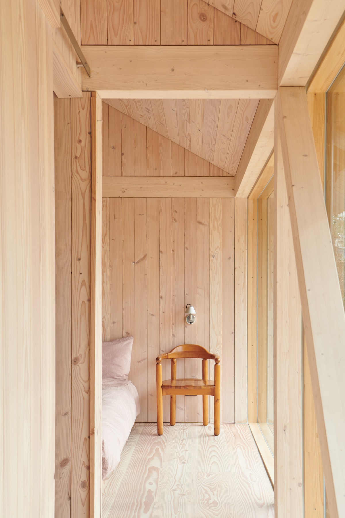 A serene sleeping area features warm wooden walls and high ceilings. A spacious bed is positioned beside a large window, allowing natural light to fill the room. A simple wooden chair rests nearby, enhancing the uncluttered and peaceful ambiance.