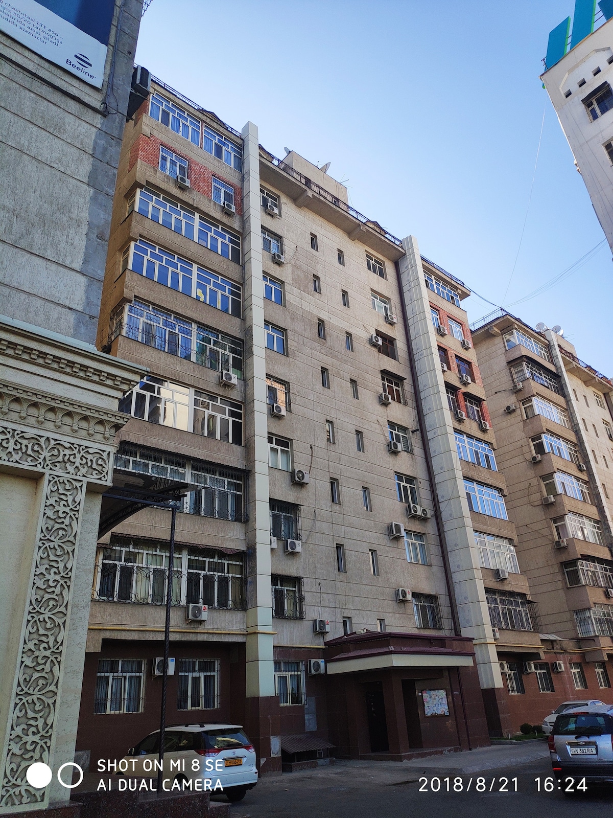 A multi-story building featuring a tan exterior with large, rectangular windows equipped with air conditioning units. The entrance is marked by a low canopy, and the surroundings include other similar structures, showcasing a mix of architectural styles.