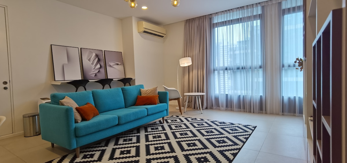 A bright living area contains a cozy turquoise sofa, complemented by decorative pillows. A patterned rug anchors the space, while large windows allow natural light to filter through sheer curtains. A stylish lamp and minimalist furniture pieces add to the modern aesthetic.