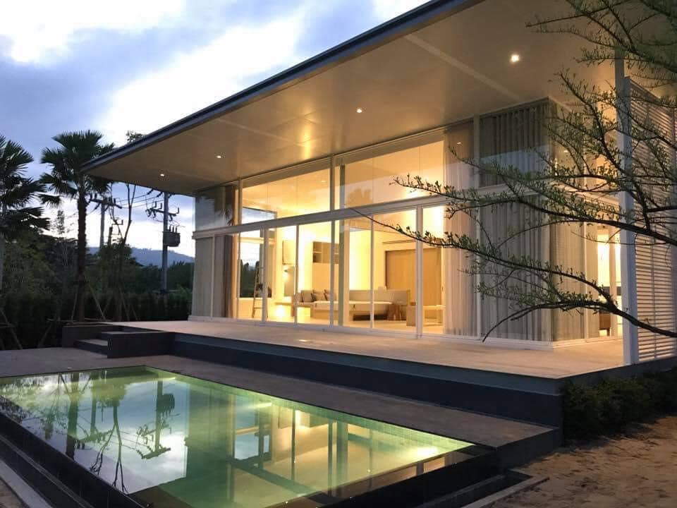 A modern house featuring large glass walls that create a seamless connection to the outdoors. A private pool sits on the wooden deck, reflecting the surrounding nature and evening sky. The landscape includes palm trees and plants, enhancing the serene environment.