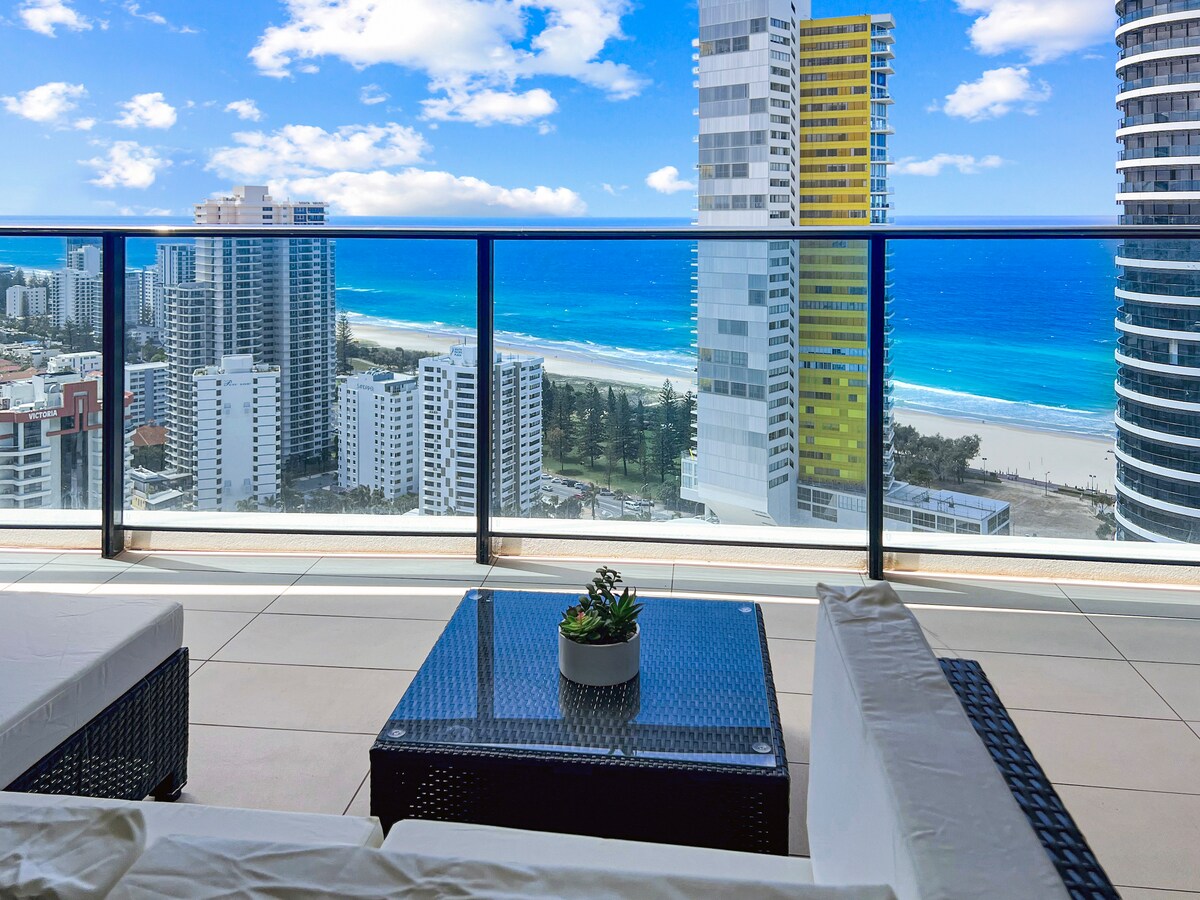 A private balcony features comfortable seating with ocean views. The expansive coastline stretches beneath a clear blue sky, with waves gently lapping at the shore. Nearby buildings reflect the vibrant colors of the Gold Coast, creating a striking snapshot of the landscape.