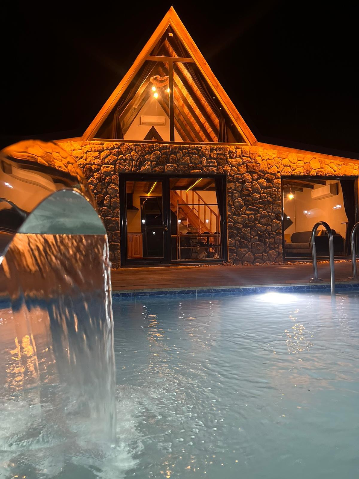 An architectural structure with a unique triangular roof is illuminated at night, showcasing warm lighting against stone walls. A sparkling heated pool reflects the light, while a fountain creates a gentle flow of water into the pool, adding a serene element.