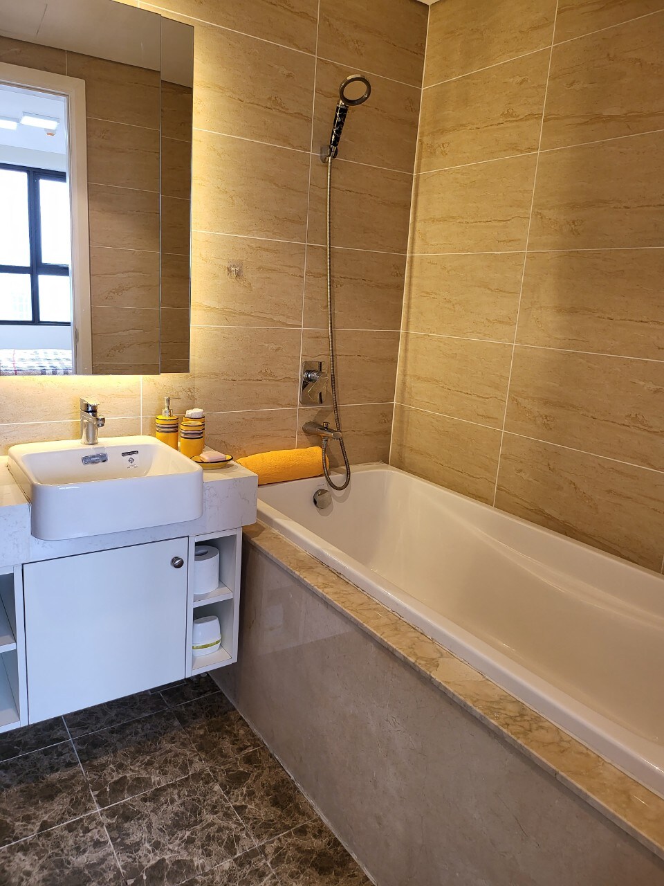 The bathroom features a modern design with beige tiled walls and a spacious bathtub. A white sink is accompanied by a mirror above, and personal care items are neatly arranged nearby. The elegant gray floor tiles add a contemporary touch to the space.
