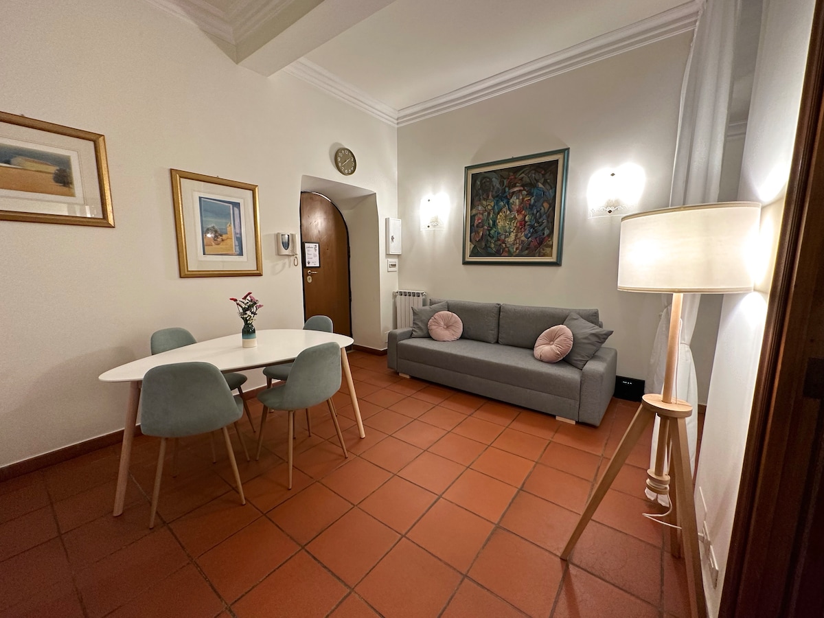 A living area features a gray sofa with decorative pillows, paired with a round dining table and four chairs. Wall art and framed pictures add visual interest. Soft lighting is provided by a standing lamp, creating a welcoming ambiance throughout the space.