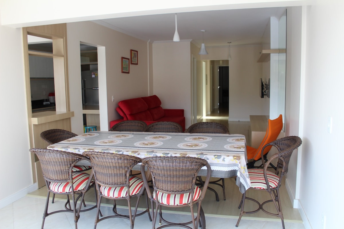 A spacious dining area is highlighted by a long table surrounded by wicker chairs with striped cushions. In the background, a living space is visible featuring a red sofa and bright lighting enhances the open layout.