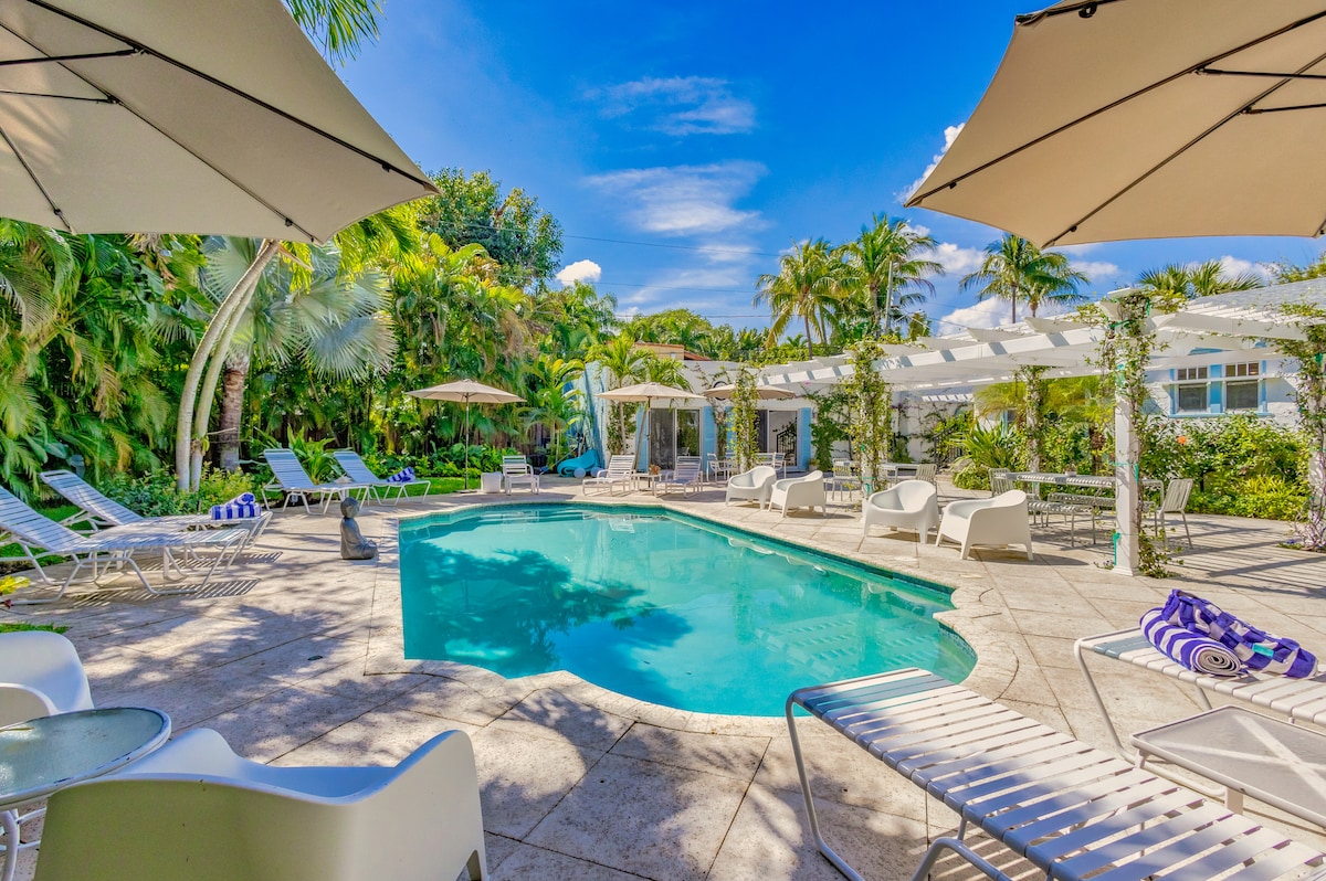 The outdoor entertainment area features a heated swimming pool surrounded by lounge chairs and shaded umbrellas. Lush tropical landscaping creates a serene environment, while a pergola enhances the space with a sense of privacy. Sunlight reflects off the water, inviting relaxation.