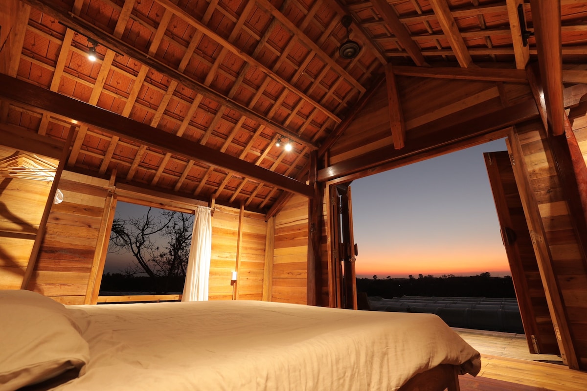 A wooden interior features a spacious bed at the center, surrounded by warm tones. Large doors open toward a serene sunset, casting gentle light across the room. Ceiling beams add character to the high, sloping roof while natural textures create a calming environment.