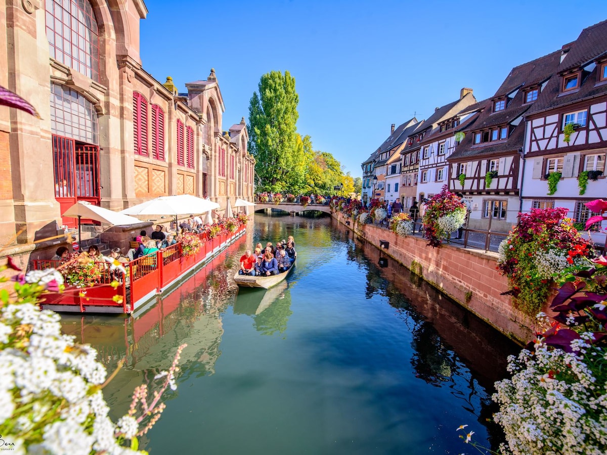 A serene canal scene is depicted, featuring charming half-timbered houses adorned with flowers. A small boat glides along the water, surrounded by lush greenery and colorful floral displays along the banks. Sunlight casts a warm glow, enhancing the quaint atmosphere.