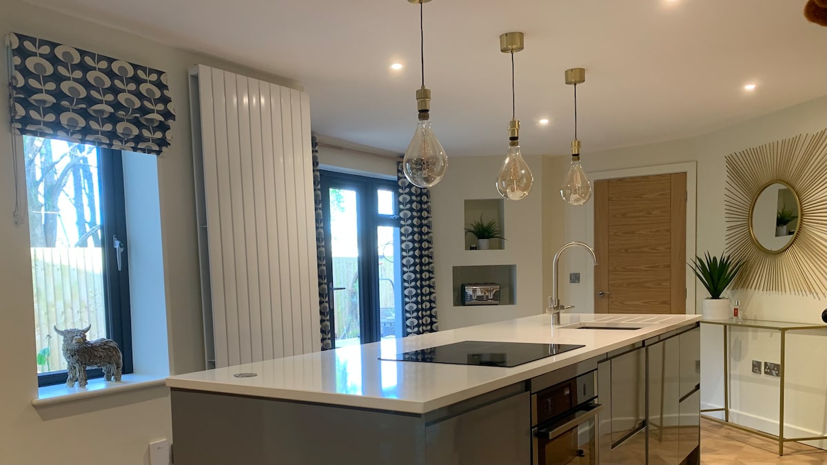 A modern open-plan kitchen features a central island with an induction cooktop. Stylish pendant lights hang above, illuminating the space. Large windows allow natural light to fill the room, highlighting the sleek cabinetry and elegant design accents, including decorative plants and patterned curtains.