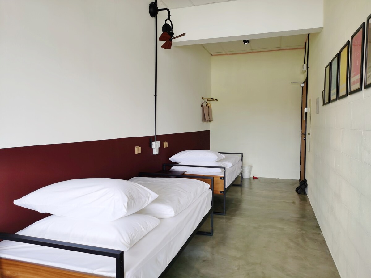 A twin bedroom features two single beds with white linens and ample pillows, positioned against a calm, burgundy accent wall. A minimalist design is evident, with a coat rack and framed artworks displayed on the adjacent wall. The concrete floor complements the modern aesthetic.