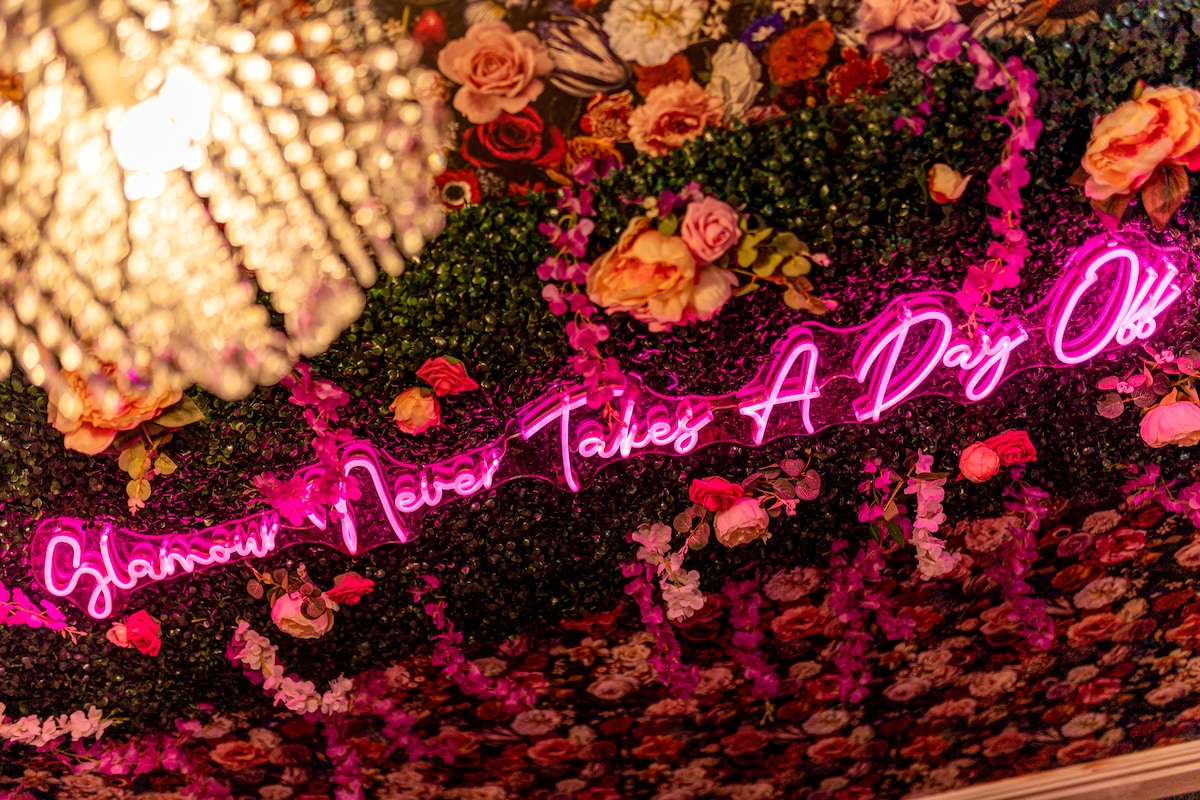 A decorative ceiling is adorned with vibrant floral arrangements, featuring a blend of greenery and multicolored roses. A neon sign reading 'Glamour Never Takes A Day Off' adds a striking element. A chandelier overhead provides soft illumination to the space.