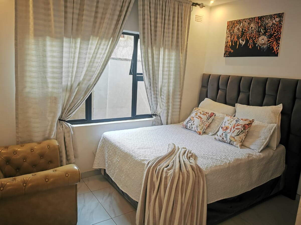 A cozy sleeping area features a spacious bed with decorative pillows and a textured white blanket. Natural light filters through a window with sheer curtains. A beige armchair adds comfort, while a colorful wall art piece adds a touch of design above the bed.
