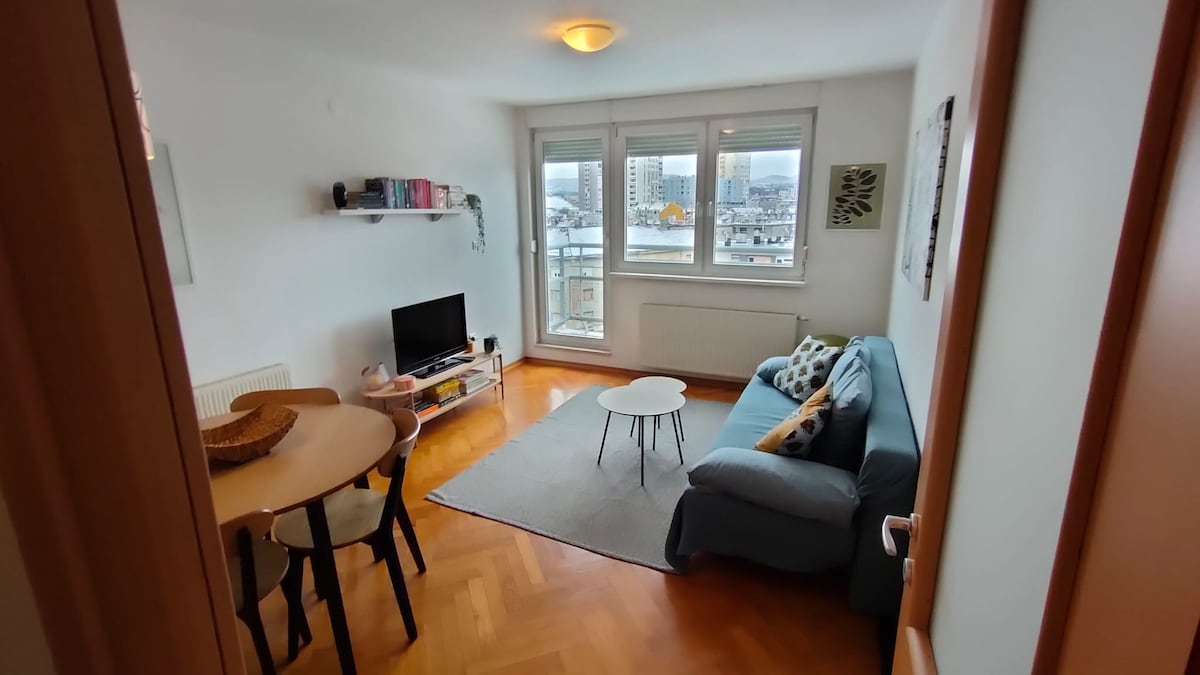 A living area features a comfortable sofa and a small round coffee table. A dining area with a wooden table and chairs is visible, alongside a wall-mounted shelf displaying books. Large windows provide natural light and offer a view of the cityscape.