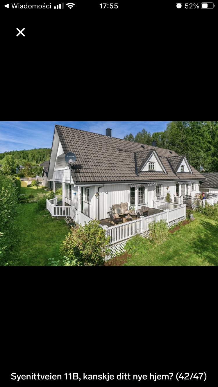 A charming two-story residence is surrounded by lush greenery, featuring white siding and a traditional pitched roof. The home includes multiple outdoor seating areas on the balcony, with a manicured garden enhancing the inviting exterior. Sunlight filters through the trees, contributing to a serene atmosphere.