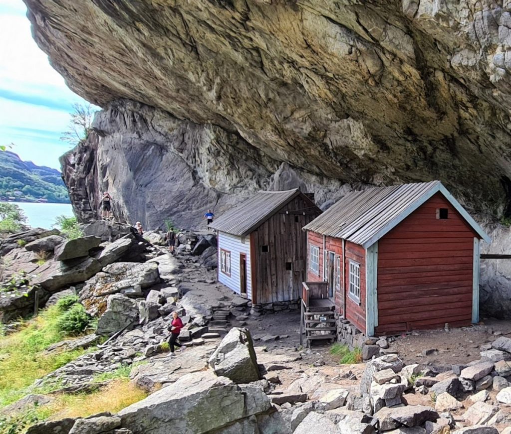 Two wooden cabins sit against a large rock overhang, surrounded by rugged terrain. A path leads towards the cabins, and a lush landscape is visible beyond. The scene showcases a blend of natural beauty and rustic architecture, with a clear view of the water in the distance.