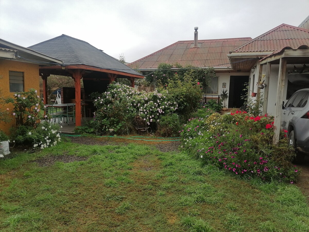 A garden area features a variety of colorful flowers and greenery, providing a serene outdoor space. A wooden gazebo stands in one corner, with a table and chairs partially visible. A separate building is seen in the background, indicating a well-maintained outdoor environment.