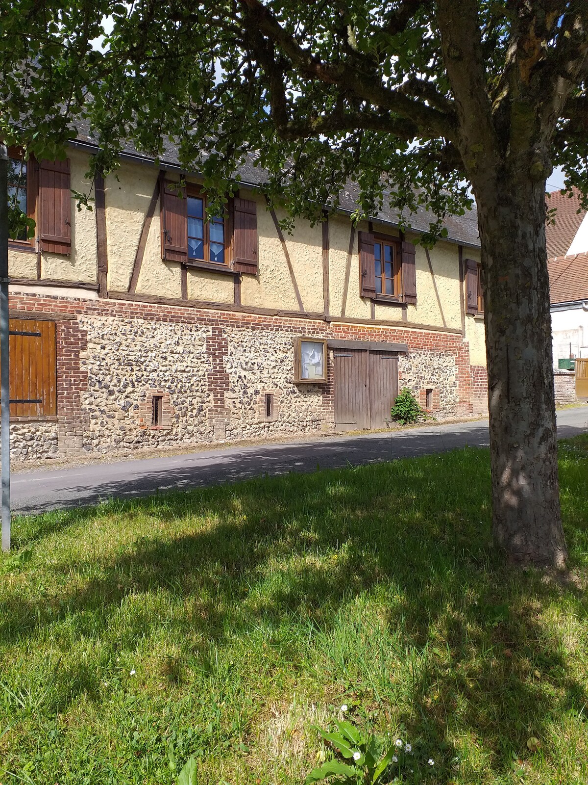 The exterior of a charming longère features a combination of stone and timber construction. Wooden shutters adorn the windows, and a rustic wooden door is visible. Grass and trees surround the building, creating a natural setting.