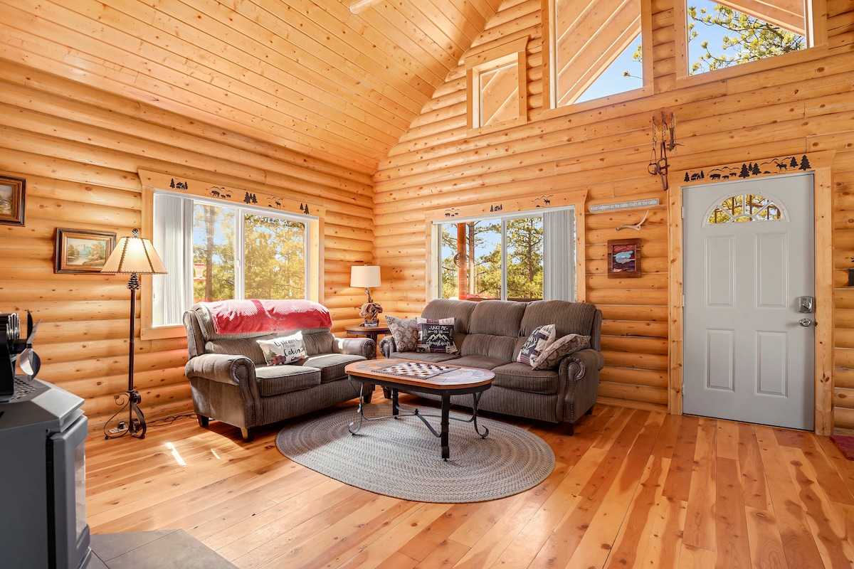 A cabin interior features log walls and high ceilings, allowing natural light to illuminate the space. Two comfortable sofas are placed around a central coffee table on a circular rug. A warm atmosphere is created by the wooden flooring and tasteful decor, enhancing the cozy setting.