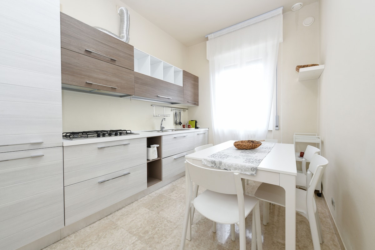 The kitchen features modern cabinetry in light and dark tones, complemented by a spacious countertop. A white dining table with four chairs is positioned near a large window, allowing natural light to illuminate the area. Essential appliances are neatly arranged for convenience.