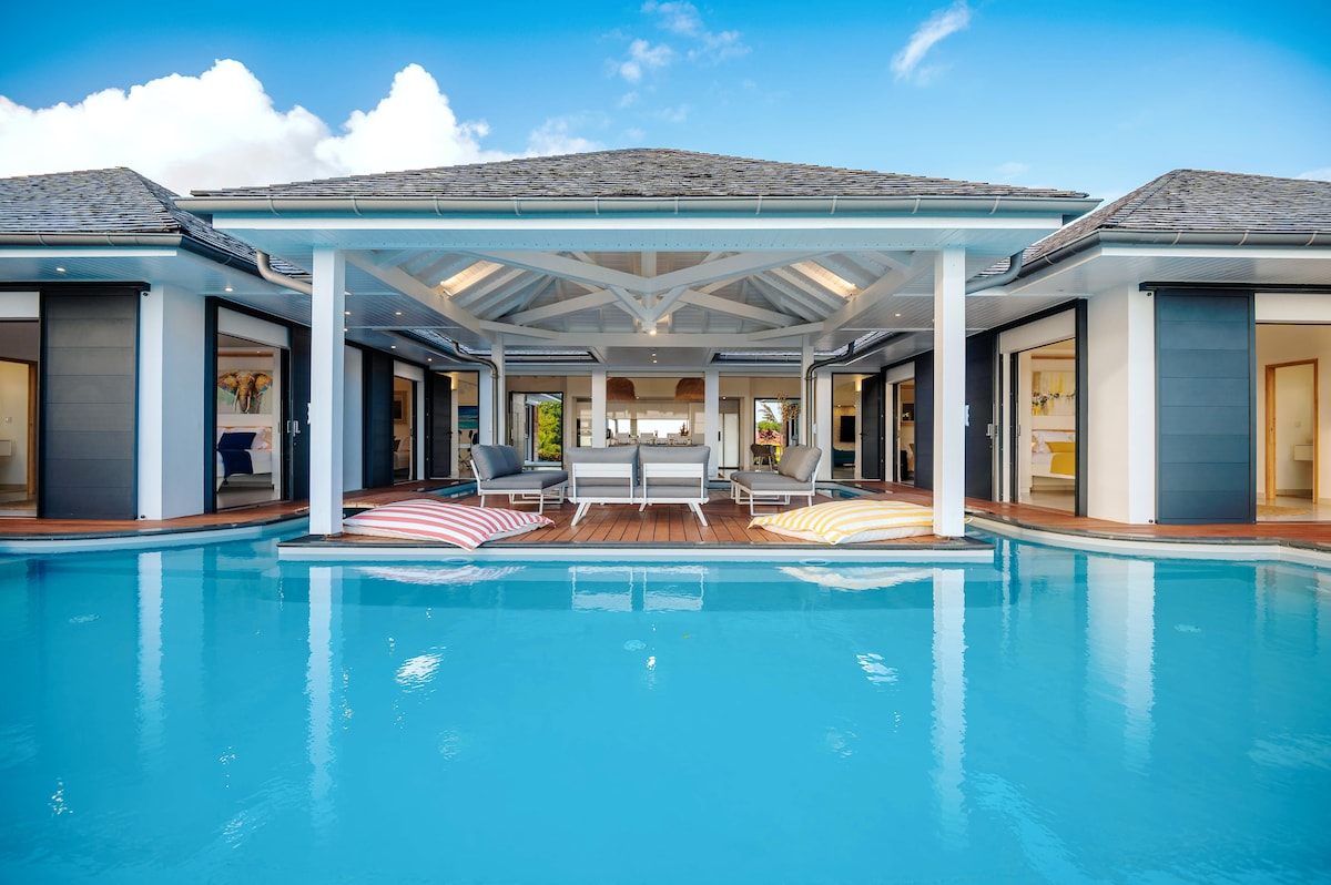 The exterior of the villa is highlighted by a spacious, inviting pool that reflects the sky. The structure features an open design with large glass doors, leading to multiple entries from the deck. Lounge chairs are positioned on the pool's edge, inviting relaxation.