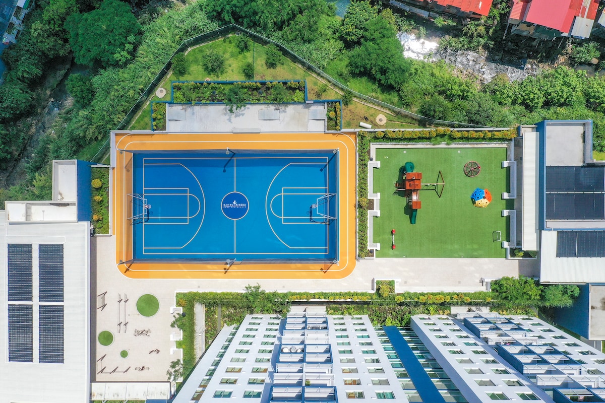 An aerial view captures a well-maintained sports court with a vibrant orange perimeter, alongside a grassy area equipped with playground structures and colorful seating. Trees and lush landscaping enhance the surroundings, providing a serene environment for outdoor activities.