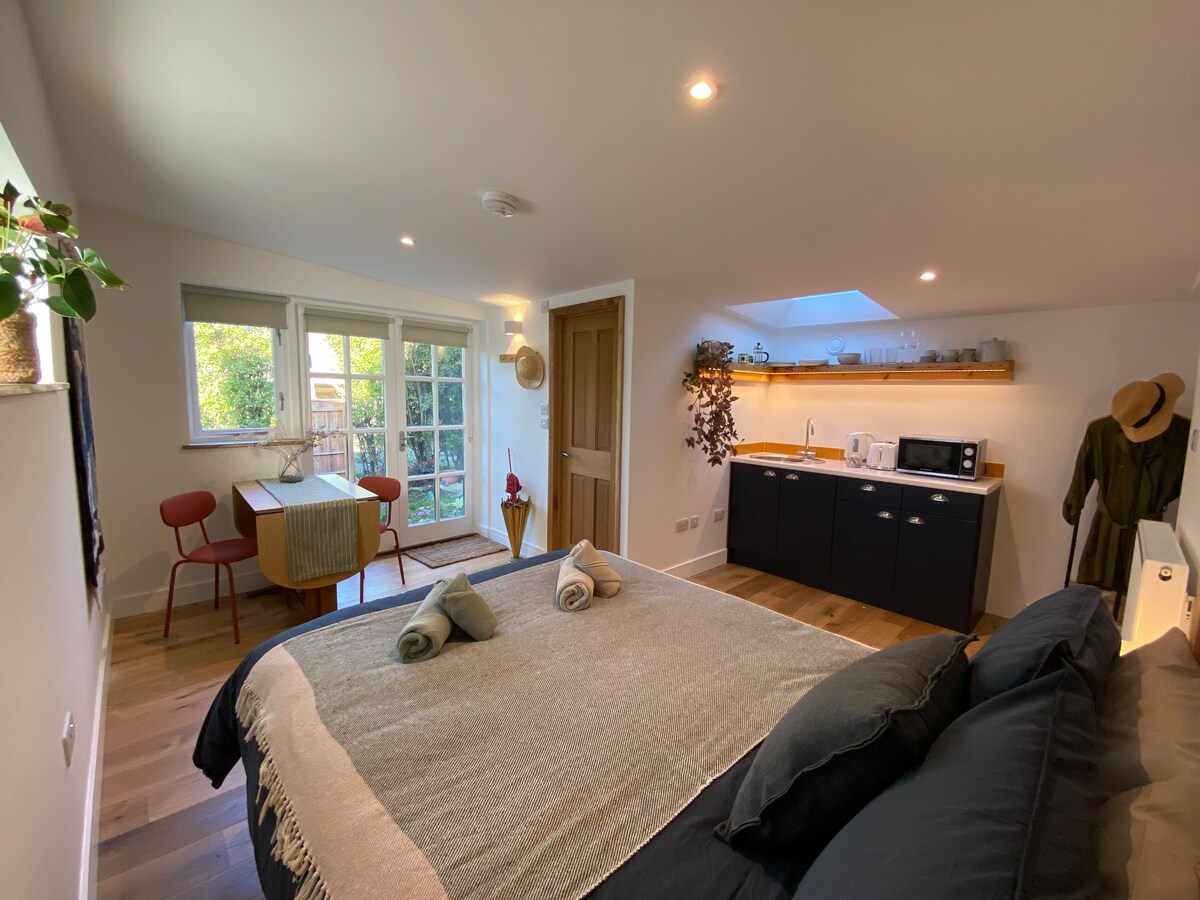 A modern studio space is presented with a spacious bed adorned with textured pillows. A dining area with two red chairs and a wooden table is positioned next to a kitchenette featuring essential appliances. Natural light enters through the windows, enhancing the inviting environment.