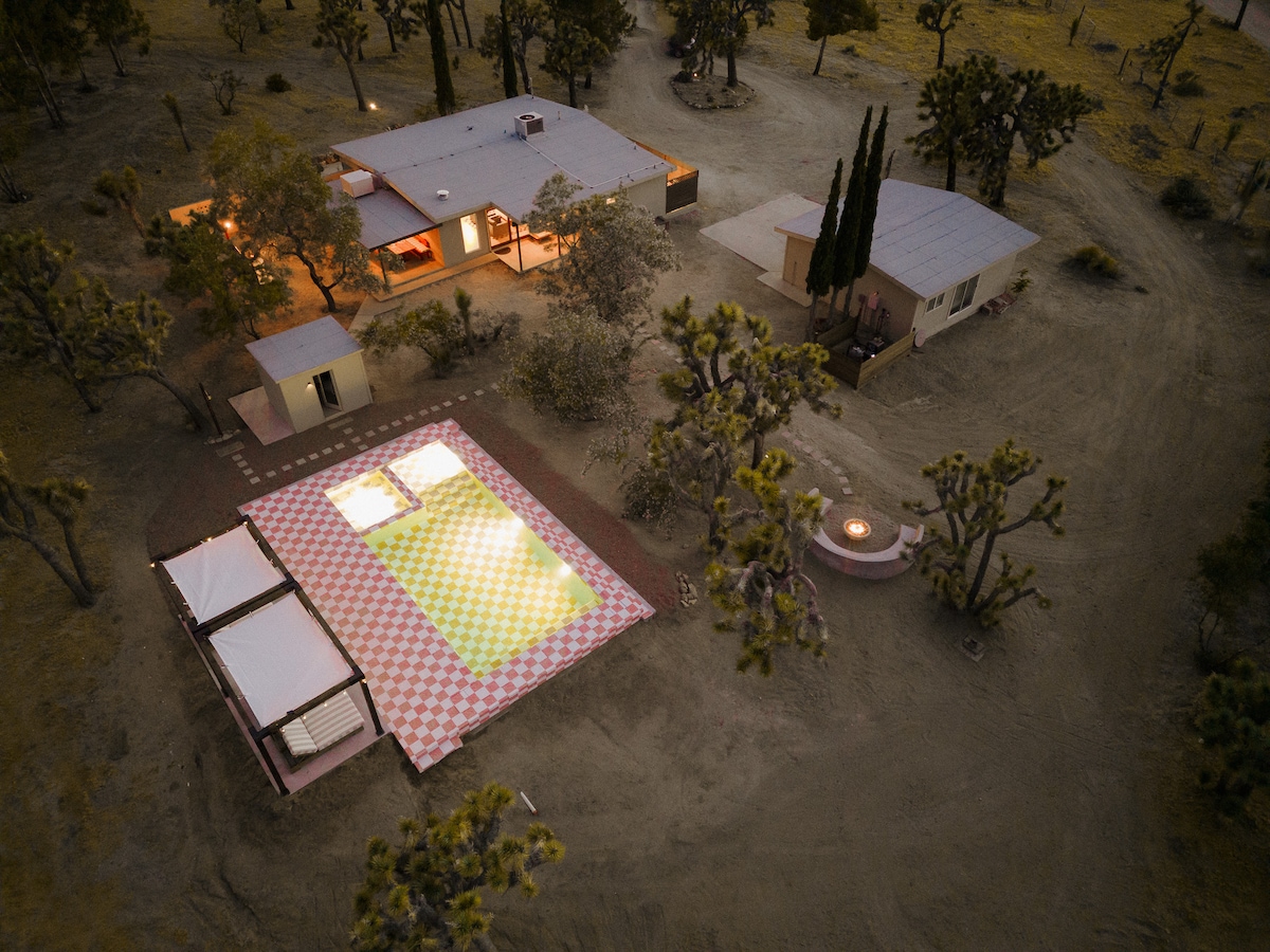 An aerial view showcases the sprawling desert property, featuring a designer pool surrounded by checkered tiles, two shaded cabanas, and a fire pit nearby. The main house and an adjacent structure are nestled among the Joshua Trees, highlighting the desert's natural landscape.