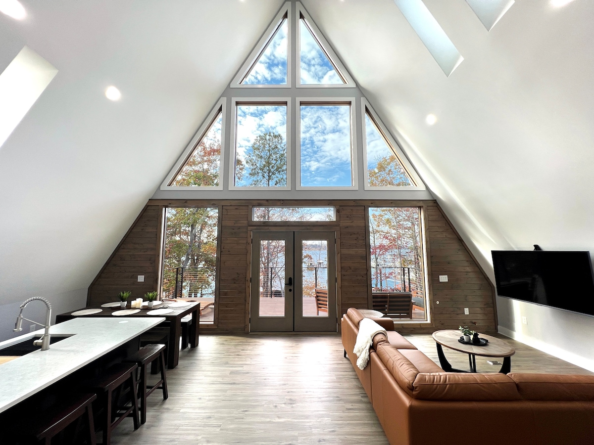 The spacious, open floor plan features large triangular windows that provide views of Lake Keowee and surrounding trees. A comfortable leather couch faces a mounted TV, while a modern dining area with a long table is visible. Light reflects off the wooden floor, enhancing the airy feel.