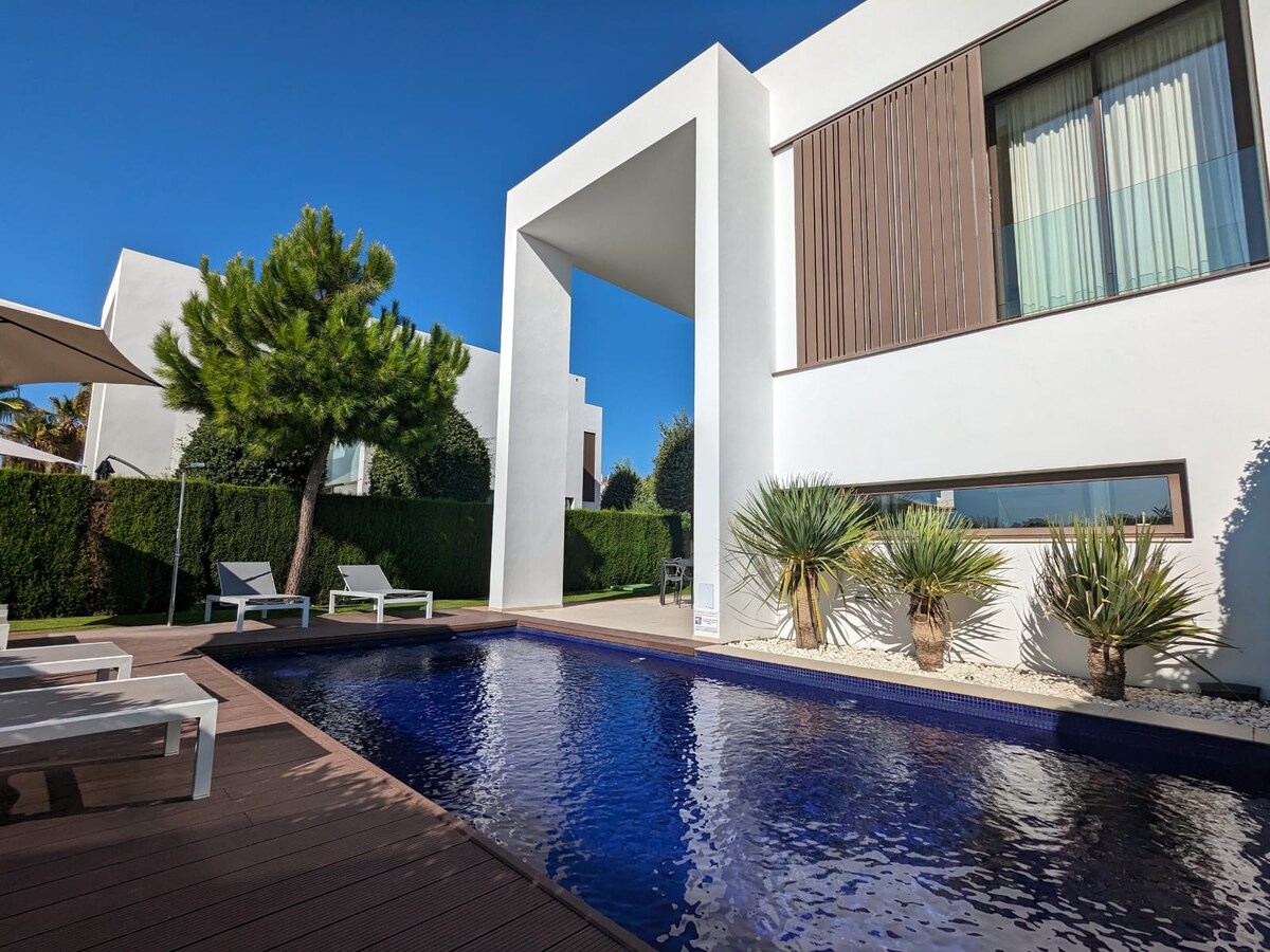A contemporary villa is featured with a private pool framed by landscaped greenery. Sun loungers line the poolside, while a large window offers a view of the serene outdoor area. The clean lines of the architecture complement the bright blue sky above.