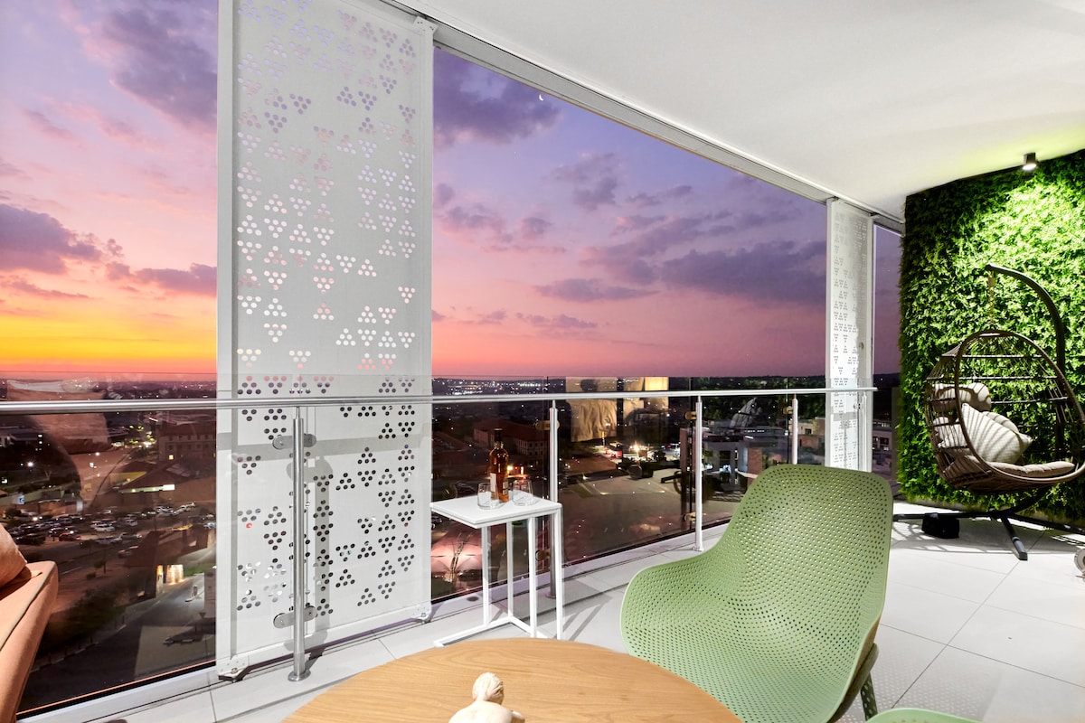 A spacious balcony features an outdoor seating area with modern green chairs and a small table. Glass partitions frame the view of a vibrant sunset over the city. The wall is adorned with greenery, adding a natural element to the space.