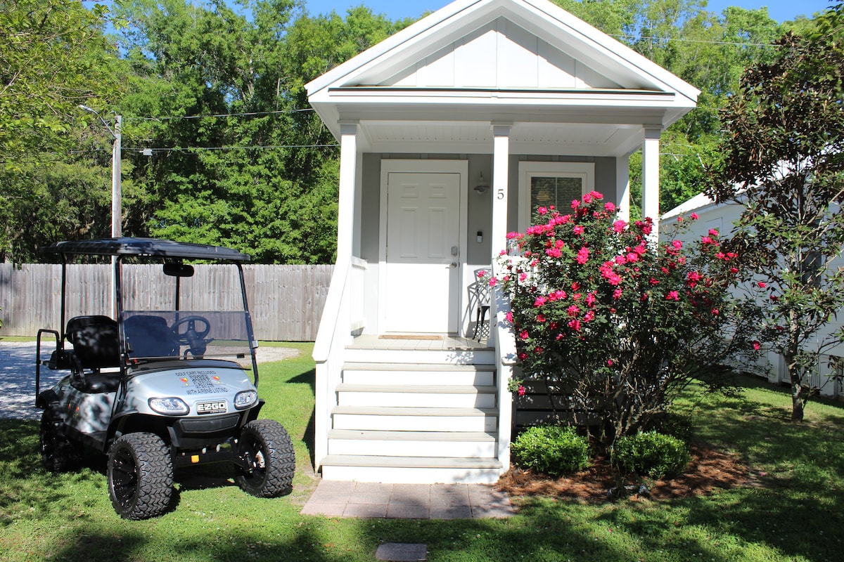 The exterior of the cottage features a welcoming front porch with two steps leading up to the entrance. A golf cart is parked nearby on a grassy area, surrounded by blooming pink flowers and green landscaping.