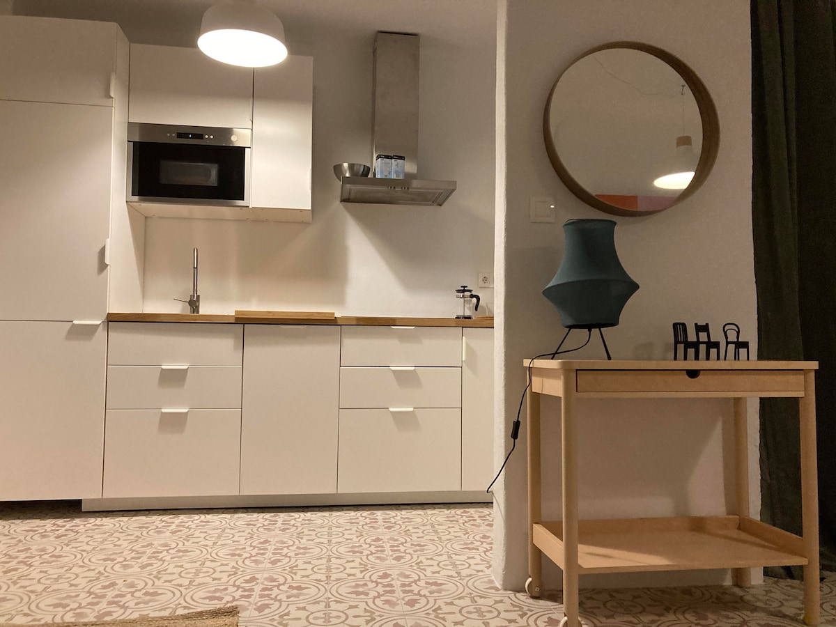 A modern kitchen area is visible, featuring sleek white cabinetry and a wood countertop. A round mirror hangs on the wall, reflecting light. The space is illuminated by a pendant light, and a small cart with a decorative lamp is placed nearby.