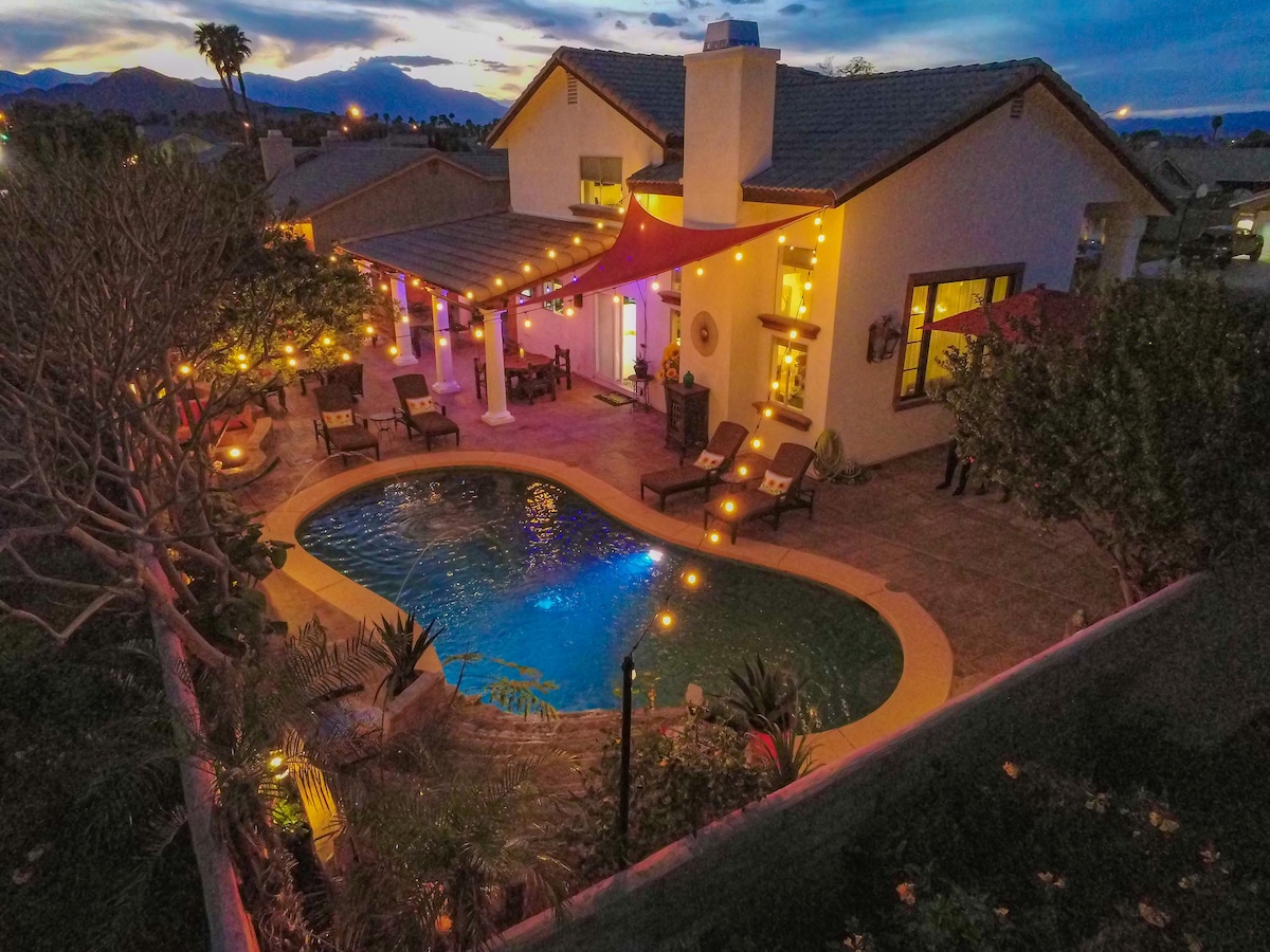 An outdoor area is captured at dusk, showcasing a pool surrounded by elegant loungers and vibrant greenery. String lights illuminate the space, creating a warm atmosphere. A covered patio provides shade, and the distant mountains are visible against a twilight sky.