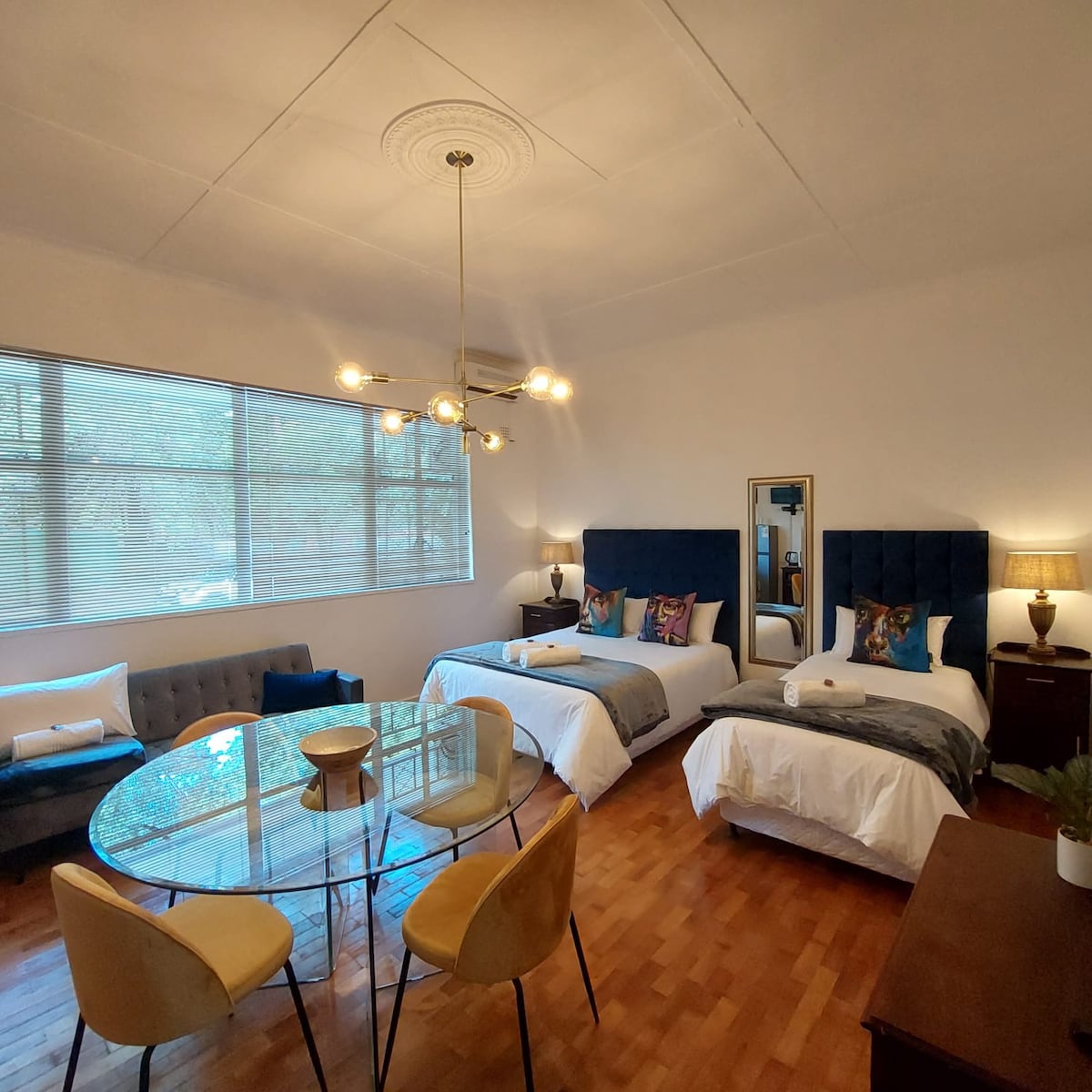 A bright bedroom features two spacious beds with blue headboards. A mirrored accent portrays a stylish décor. A glass dining table with four yellow chairs occupies the center, while a sofa and window offer natural light, enhancing the open atmosphere.