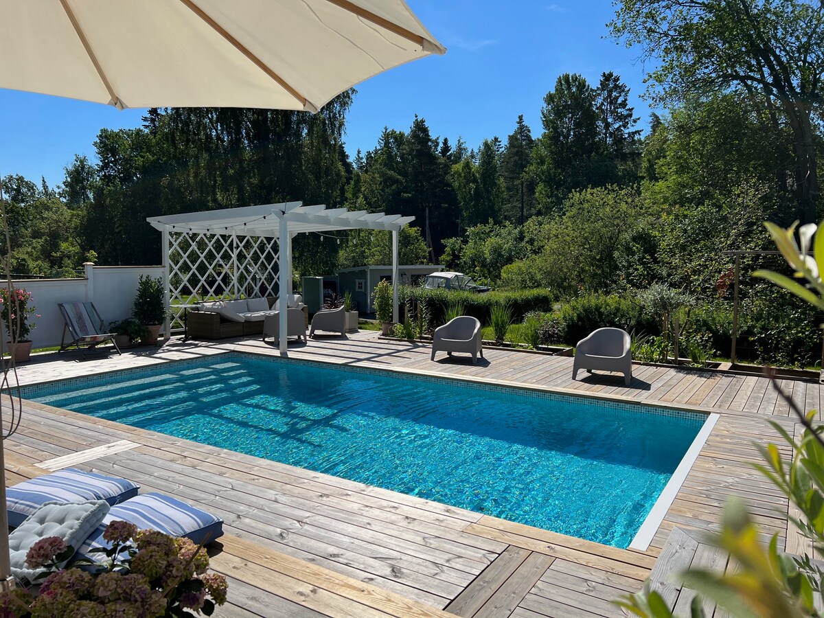 A spacious outdoor area features a rectangular pool with clear blue water, surrounded by a wooden deck. Comfortable lounge chairs are positioned around the pool, and a shaded pergola provides a cozy seating area. Lush greenery enhances the tranquil setting.