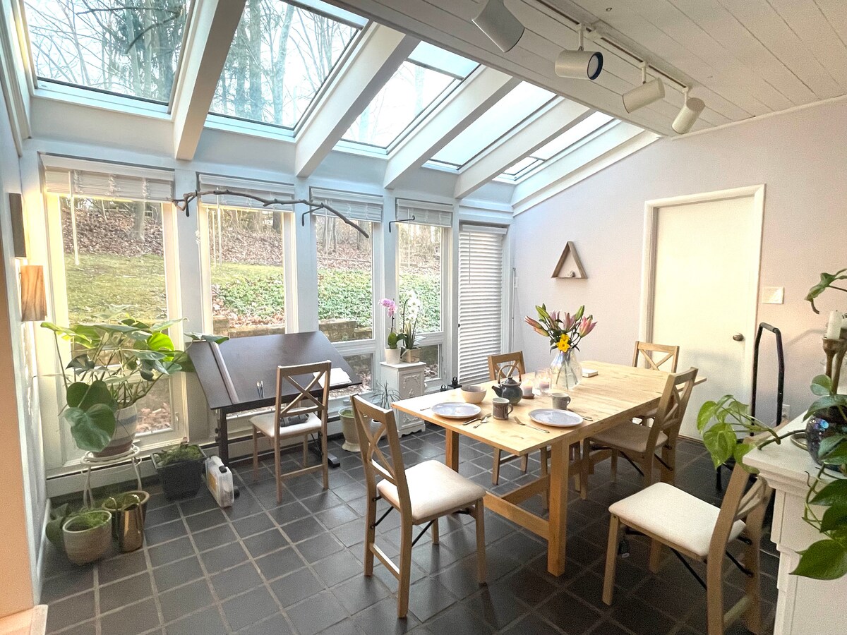 A sunlit dining area features large skylights and floor-to-ceiling windows, providing a view of the surrounding greenery. A wooden table surrounded by six chairs is set with tableware. Potted plants add a touch of nature to the space, enhancing the light, airy atmosphere.
