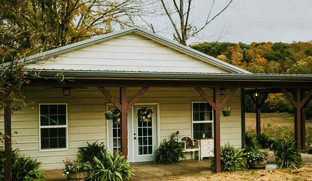 The exterior of a charming cottage is depicted, showcasing a covered porch with wooden beams. Lush greenery and decorative plants frame the entrance, while large windows allow for natural light. The surrounding landscape features autumn-colored trees, enhancing the tranquil setting.
