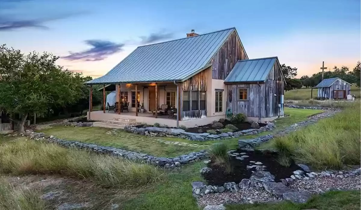 A rustic home with a metal roof and wooden siding is set amidst an expansive grassy landscape. Stone pathways lead to the inviting front porch, furnished with seating. The surrounding area features trees and shrubs, contributing to the serene rural atmosphere.