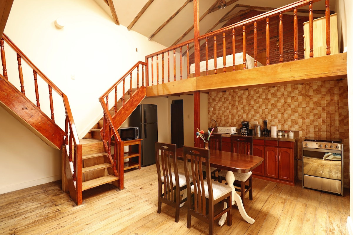 An open layout presents a spacious living and dining area featuring a wooden table with chairs. A kitchen area is visible, equipped with essential appliances. Stairs lead to an upper level with a lofted sleeping space. Natural light enhances the warm wooden tones throughout.