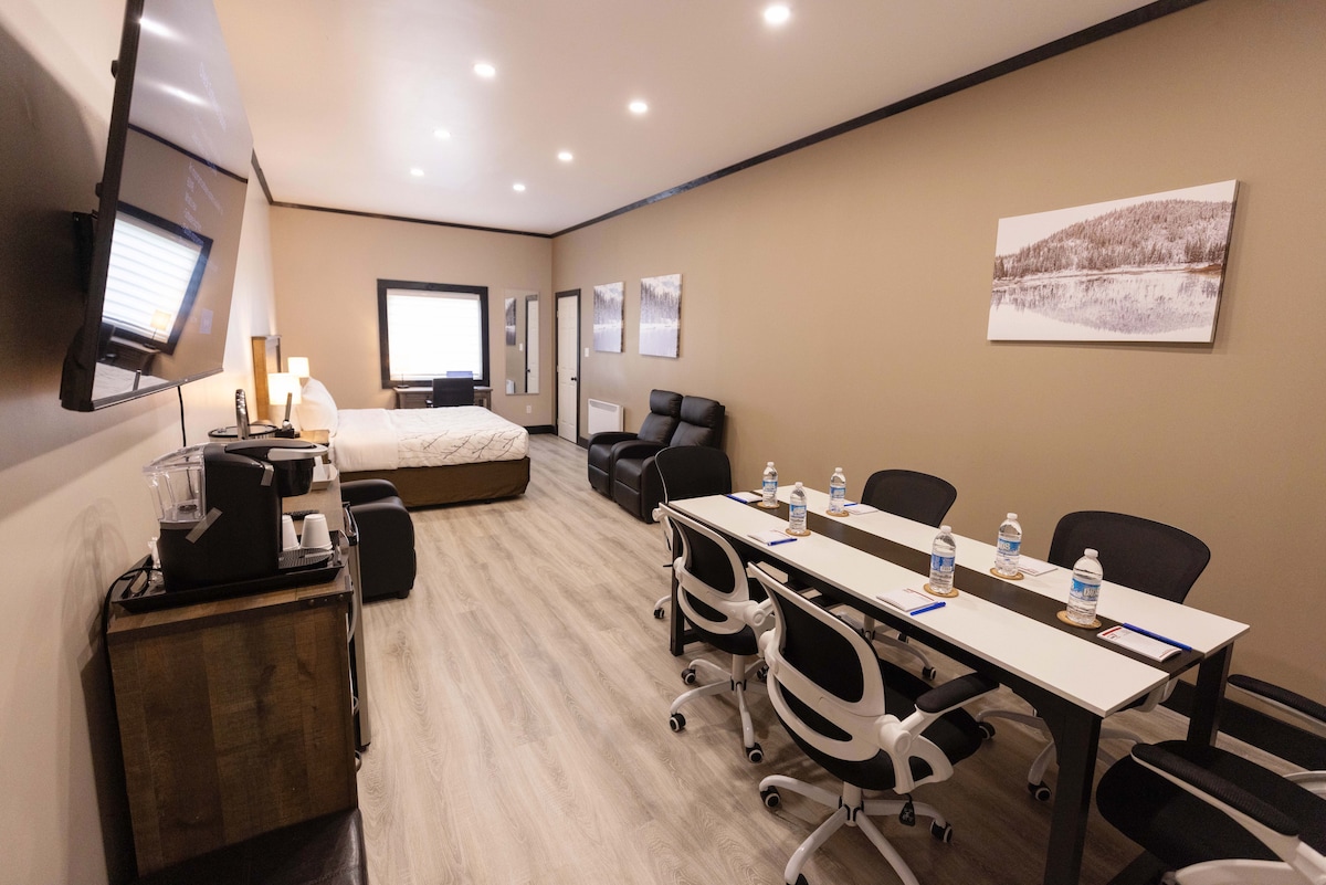 A spacious room is displayed, featuring a comfortable sleeping area with a bed and a large window. A modern workspace is arranged with a long table and several ergonomic chairs. A coffee station and a seating area with black recliners are also visible.