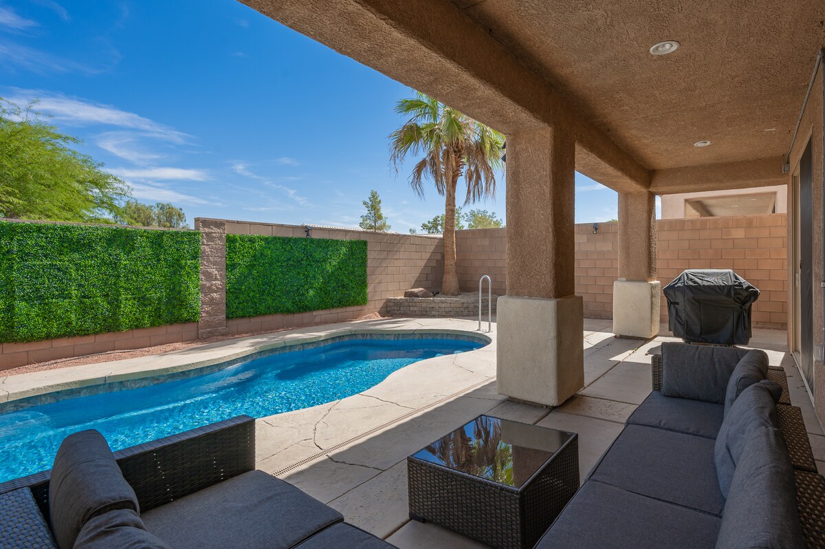 An outdoor seating area showcases a dark wicker sofa set with plush cushions, positioned next to a sparkling pool. Surrounding greenery provides privacy, while a BBQ grill sits in the corner. The sky is bright with a few clouds, enhancing the relaxed atmosphere.