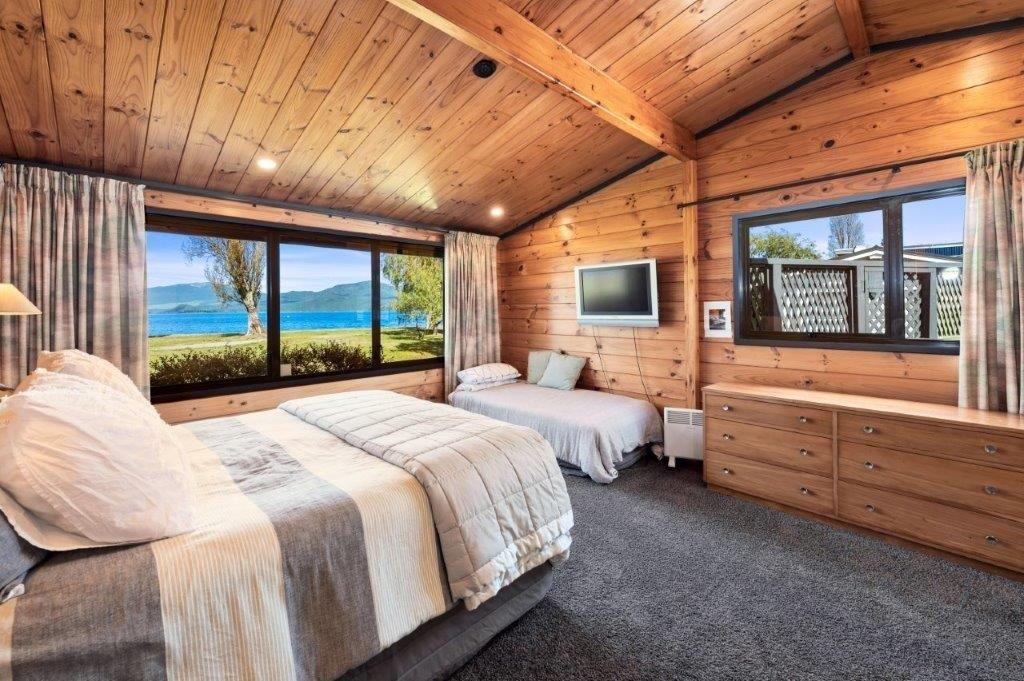 A spacious bedroom features wooden paneling and high ceilings, offering views of the lake from large windows. A comfortable bed is complemented by a cozy twin bed, while a dresser provides additional storage. Natural light brightens the room, creating a serene ambiance.