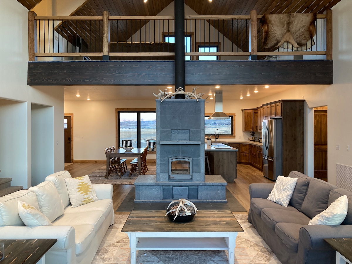 An open-concept living area features a central stone fireplace, surrounded by comfortable seating. Large windows offer views of the outdoors, while a dining set is visible in the background. A functional kitchen with dark wooden cabinetry and modern appliances is seamlessly integrated into the space.