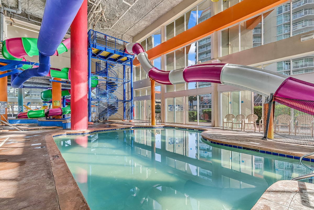 An indoor water park area features colorful water slides in various shapes and sizes, surrounded by a clear swimming pool. Floor-to-ceiling windows allow natural light to fill the space, highlighting the playful atmosphere designed for family enjoyment.