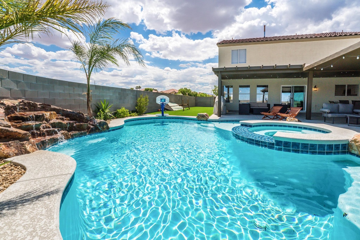 A private backyard features a sparkling pool with a waterfall and integrated hot tub. Lounge chairs are positioned nearby under a covered patio, with green grass and palm trees visible in the background. The sky is bright with scattered clouds, adding to the tranquil ambiance.
