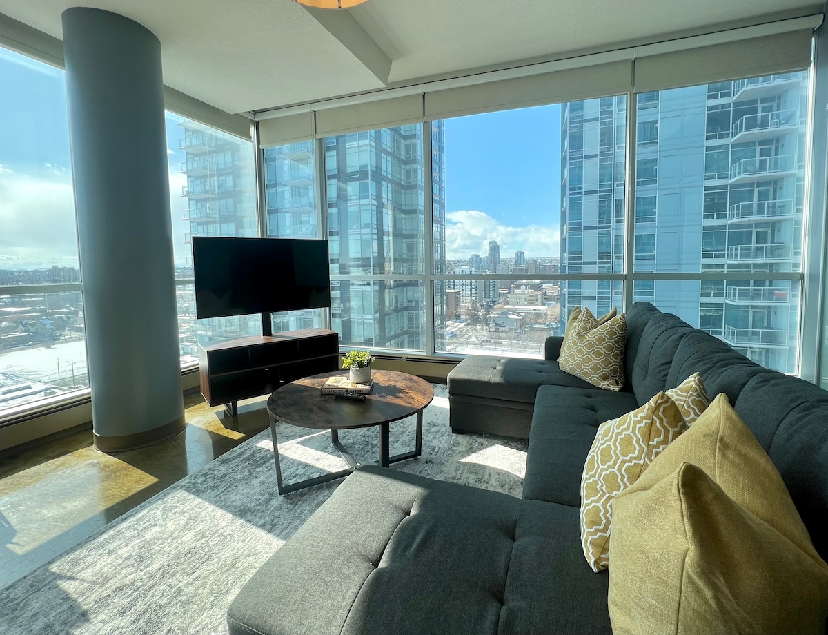 A modern living area features a spacious sectional sofa adorned with patterned cushions, facing a flat-screen TV. Sunlight floods through large floor-to-ceiling windows, highlighting the cityscape beyond. A round coffee table rests atop a soft area rug, enhancing the welcoming ambiance.
