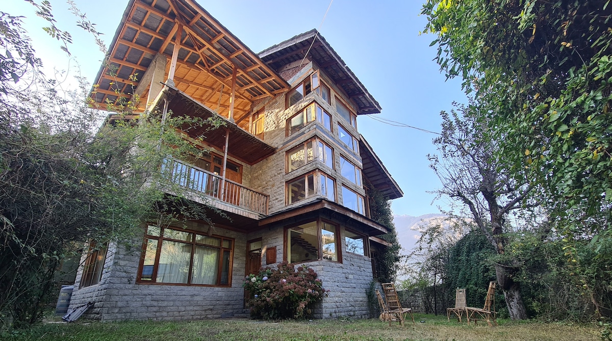 The multi-level villa features a stone and wooden exterior, framed by large windows that provide views of the surrounding greenery. A well-maintained garden area includes wooden chairs for outdoor relaxation, enhancing the connection with nature.