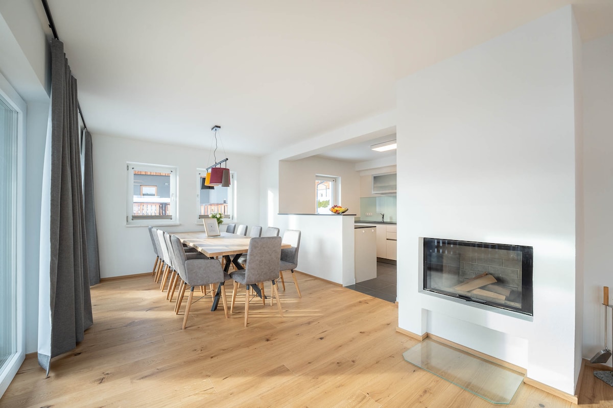An open dining area features a large wooden table surrounded by gray upholstered chairs. A modern fireplace is positioned within a white wall, complementing the minimalist design. Natural light is welcomed through nearby windows, creating a bright and airy environment.