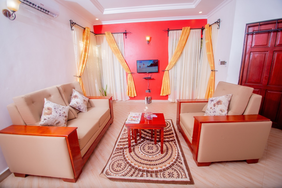 A comfortable seating area is presented with two beige armchairs and a matching sofa, featuring decorative cushions. A small red coffee table sits atop a patterned rug. Sheer curtains frame large windows, allowing natural light to brighten the space.