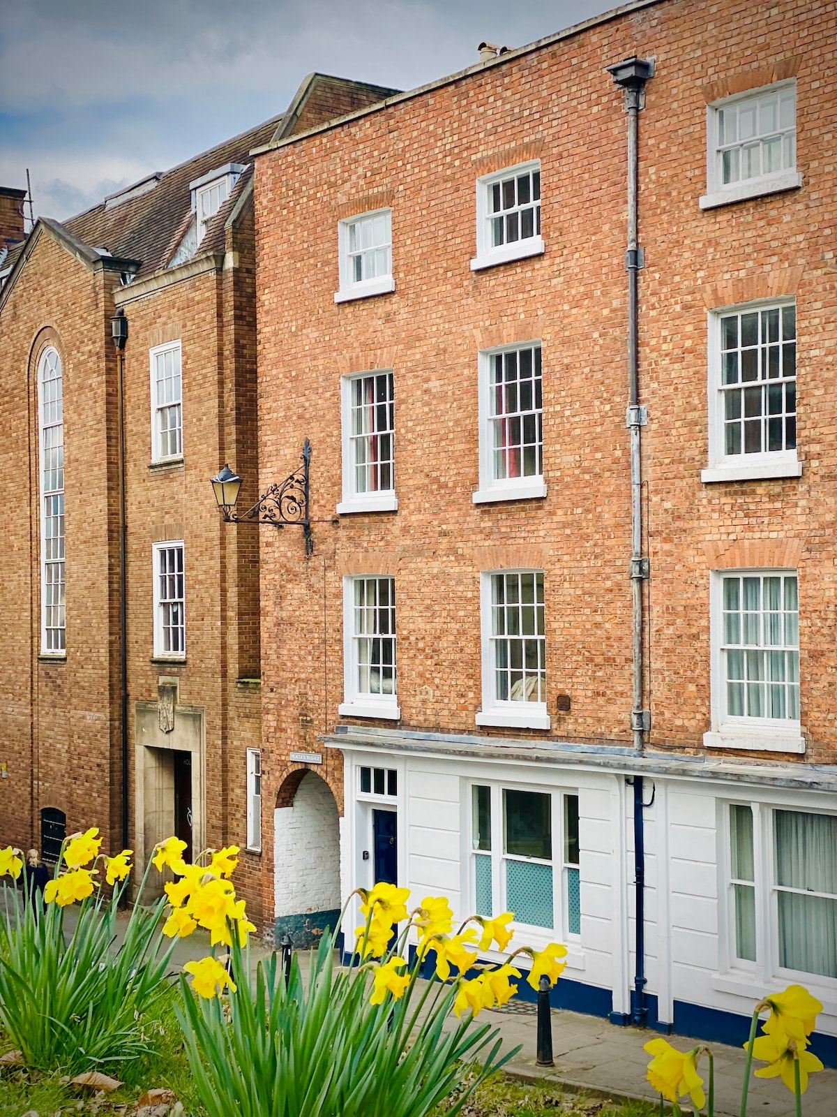 The exterior of a Georgian townhouse is shown, featuring a traditional blue front door and a light brick facade. Flowering daffodils in front add a touch of color, while large windows reflect the surrounding light.